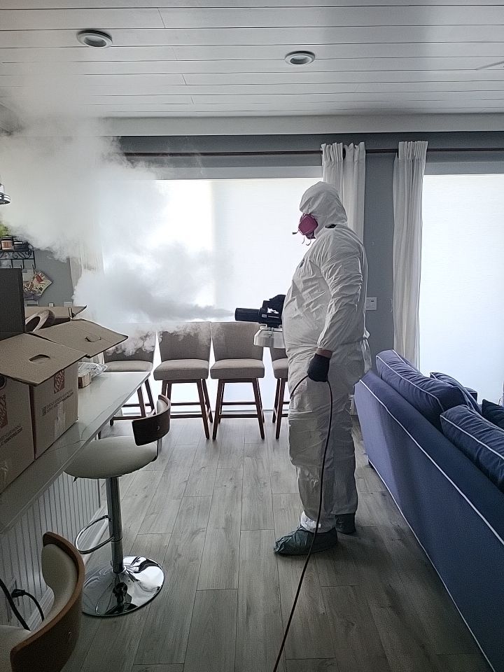 Technician in full PPE thermal fogging living room with moving boxes staged